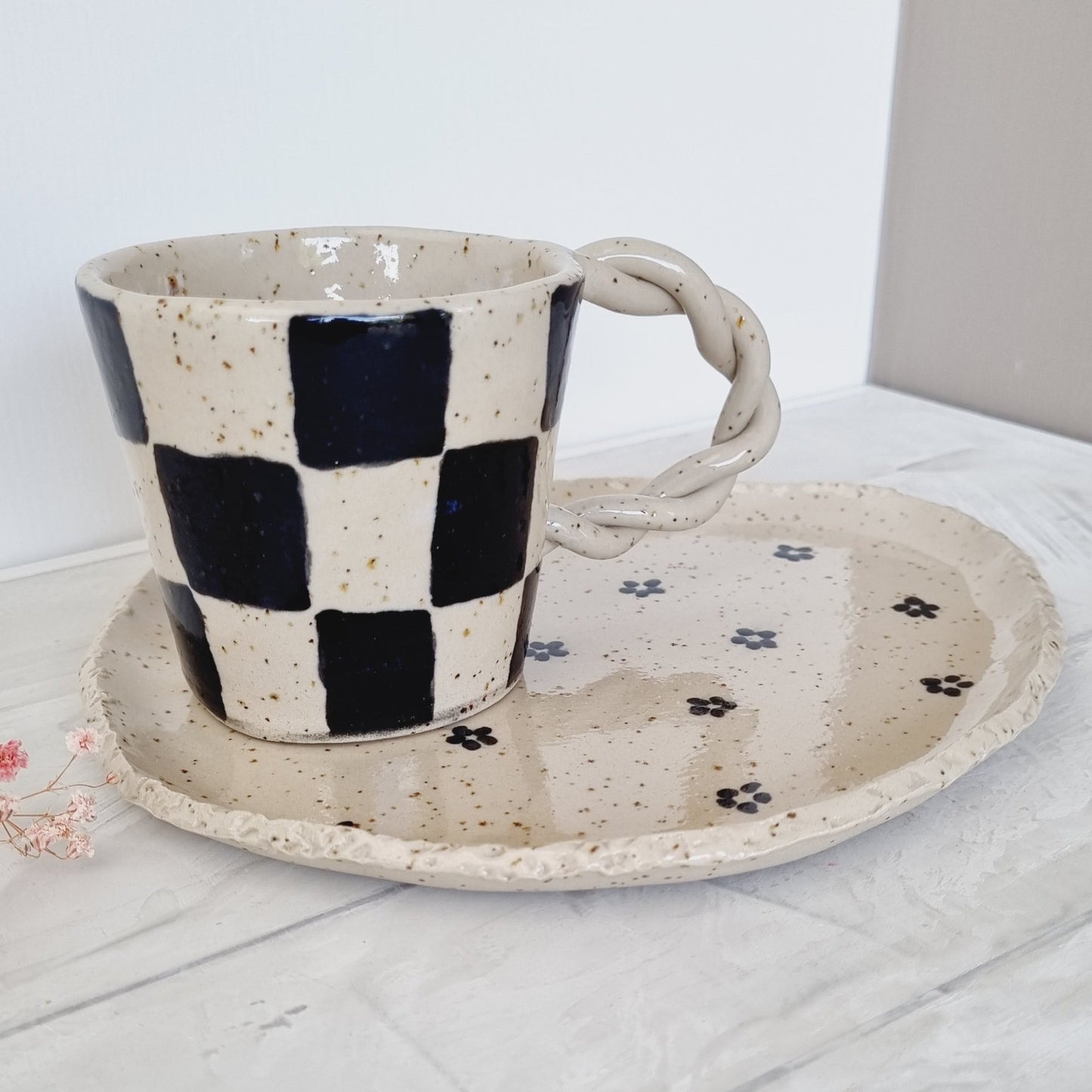 Breakfast set made from speckled clay. Mug has a black checkers pattern. Plate has an organic form nd is handpainted with black daisies