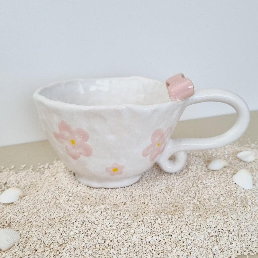 White handmade coffeecup with pink ceramic bow on the rim. Handpainted with pink flowers. Curly vintage style handle.